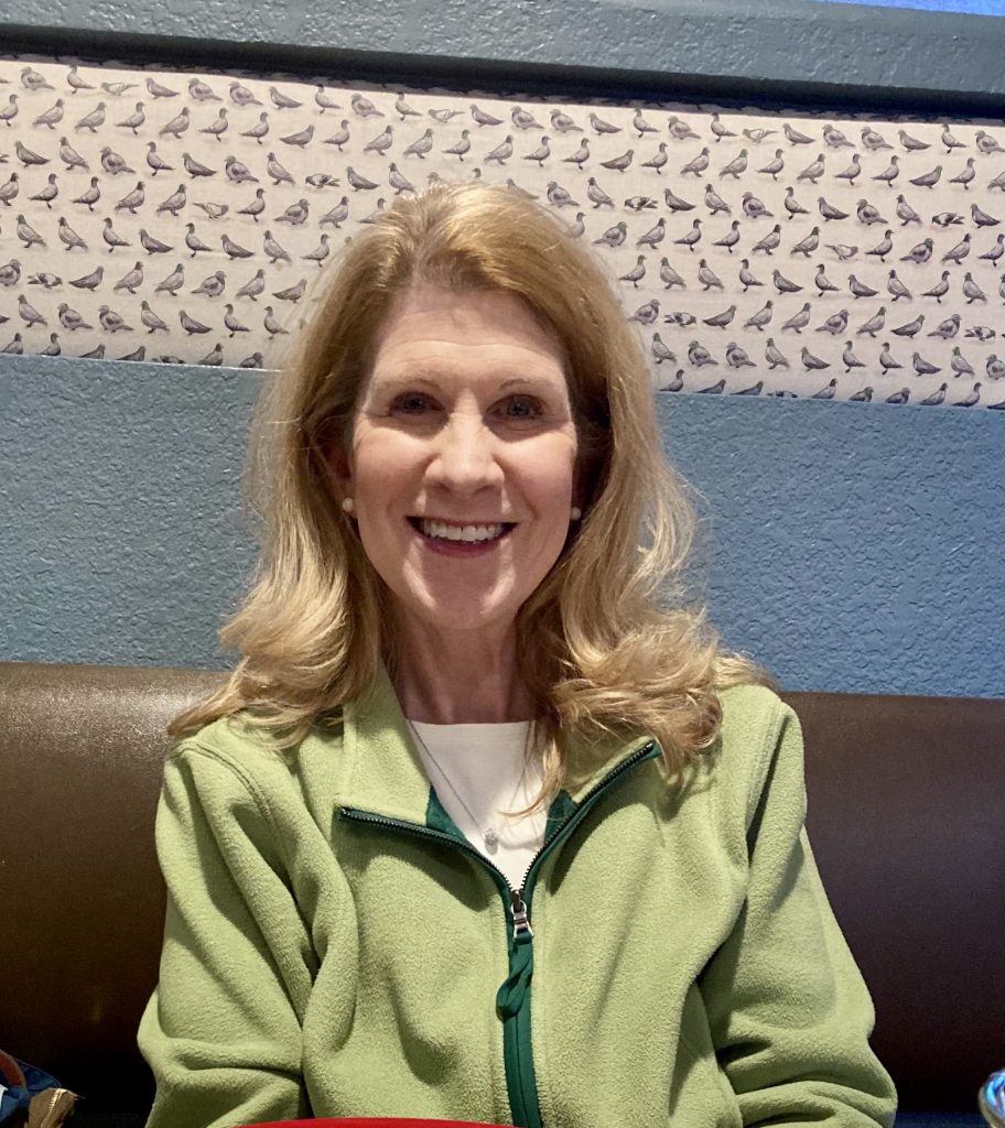 Smiling woman in green jacket with bird-patterned wallpaper.